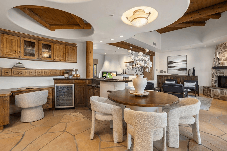 An expansive, rustic-modern open-concept living space with flagstone floors, featuring a kitchen with wood cabinets, a dining area with a round table and bouclé chairs, and a living room with a stone fireplace.