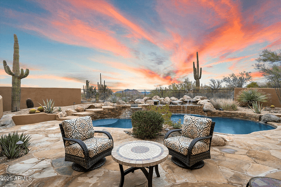 A luxurious desert backyard featuring a swimming pool with a rock waterfall, a stone patio with two chairs and a table, and saguaro cacti under a vibrant sunset sky.